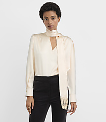 A woman wearing a cream-colored satin blouse with a neck scarf and black trousers stands against a plain white background. carousel Product Image 2