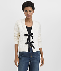 A woman wearing a white cable-knit cardigan with a black bow and blue jeans stands against a plain white background. carousel Product Image 1