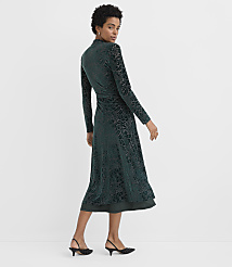 A woman wearing a long-sleeved, dark green velvet floral dress and black heels stands against a plain white background. carousel Product Image 3