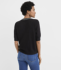 A woman wearing a black short-sleeved sweater with a beaded collar and blue jeans stands against a plain white background. carousel Product Image 3