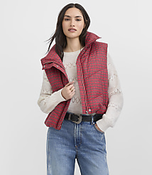 A woman wearing a red plaid puffer vest, a cream sweater, and blue jeans stands against a plain white background. carousel Product Image 2