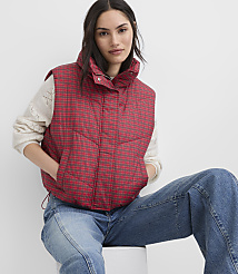 A woman wearing a red plaid puffer vest, a white sweater, and blue jeans sits against a plain white background. carousel Product Image 1