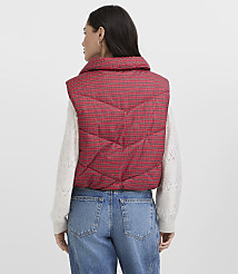 A woman wearing a red plaid puffer vest, a white sweater, and blue jeans stands against a plain light gray background. carousel Product Image 3