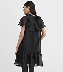 A woman wearing a black sheer dress with a large bow on the back stands against a plain white background. carousel Product Image 4