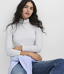 A woman with long, wavy dark hair wearing a grey turtleneck and blue jeans sits against a plain white background. carousel Product Image 2