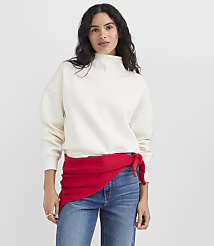 A woman wearing a white turtleneck sweatshirt and blue jeans with a red sweater tied around her waist stands against a plain light gray background. carousel Product Image 1