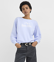 A woman wearing a light blue "Apr&egrave;s Vibes" sweatshirt and dark corduroy pants stands against a plain white background. carousel Product Image 2
