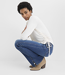 A woman wearing a white long-sleeved top, blue jeans, and tan boots crouches against a plain white background. carousel Product Image 1