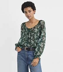 A woman wearing a green patterned blouse and blue jeans stands against a plain white background. carousel Product Image 1