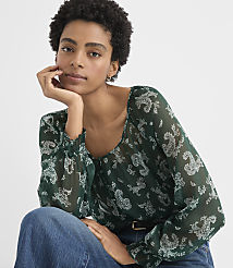 A woman with curly hair wearing a green patterned blouse and blue jeans poses against a plain white background. carousel Product Image 2