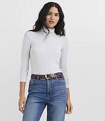A woman wearing a grey turtleneck and blue jeans with a brown belt stands against a plain white background. carousel Product Image 1
