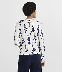 A woman with curly hair wearing a white floral sweater and dark blue pants stands against a plain light gray background. carousel Product Image 3