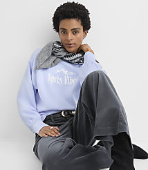 A woman wearing a light blue sweatshirt, a grey scarf, and dark corduroy pants sits against a plain white background. carousel Product Image 1