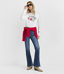 A woman wearing a white graphic long-sleeve shirt, blue flare jeans, and a red sweater tied around her waist stands against a plain white background. carousel Product Image 3