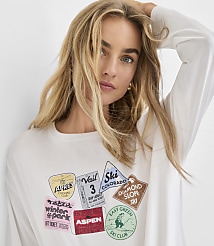 A blonde woman wearing a white long-sleeved shirt with colorful ski-themed patches poses against a plain white background. carousel Product Image 1