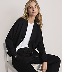 A blonde woman wearing a black cardigan over a white t-shirt and black pants sits on a white chair against a plain light-colored background. carousel Product Image 1