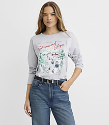 A woman wearing a grey graphic long-sleeve shirt and blue jeans stands against a plain white background. carousel Product Image 2