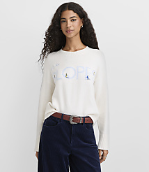 A woman with long curly hair wearing a white graphic sweater and navy corduroy pants stands against a plain light gray background. carousel Product Image 2
