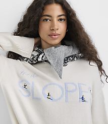 A woman with long curly hair wearing a white graphic sweater and a grey patterned scarf poses against a plain white background. carousel Product Image 1