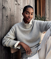A woman wearing a grey sweater and white collared shirt sits against a rustic wooden wall. carousel Product Image 4