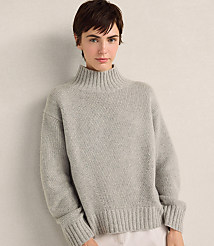 A woman with short brown hair wearing a grey ribbed turtleneck sweater stands against a plain, light-colored wall. carousel Product Image 1