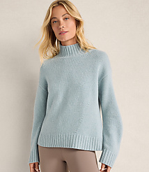 A blonde woman wearing a light blue turtleneck sweater and taupe pants stands against a plain, neutral-toned wall. carousel Product Image 1