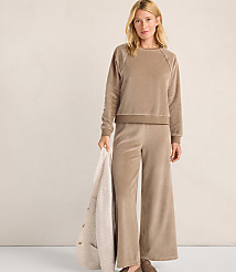 A woman wearing a tan velvet lounge set stands against a plain, light-colored wall while holding a cream-colored blanket. carousel Product Image 1