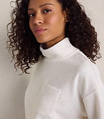 A woman with curly dark hair wearing a white textured turtleneck sweater stands against a plain, light-colored wall. carousel Product Image 3