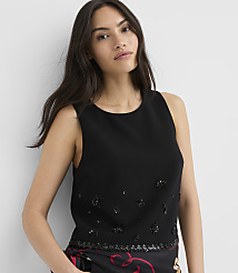 A woman with long brown hair wearing a black sleeveless top with sequin detailing stands against a plain white wall. carousel Product Image 1