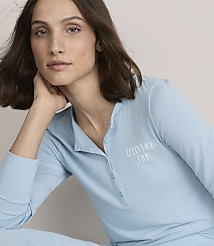 A woman with brown hair wearing light blue pajamas rests her chin on her hand against a plain, neutral background.