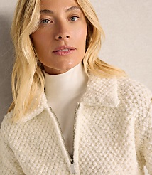 A blonde woman wearing a white turtleneck and a textured white jacket poses against a plain, light-colored wall. carousel Product Image 3