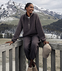 A smiling woman in a charcoal fleece set sits on a wooden railing against a backdrop of snow-capped mountains and evergreen forests. carousel Product Image 5