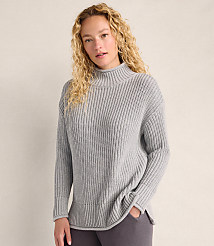 A woman with curly blonde hair wearing a grey ribbed sweater stands against a plain, light-colored wall. carousel Product Image 1