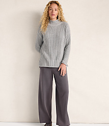 A woman with curly blonde hair wearing a grey ribbed sweater and wide-leg charcoal trousers stands against a plain, light-colored wall. carousel Product Image 2