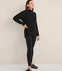 A woman wearing a black ribbed sweater, black leggings, and grey slippers stands against a plain, light-colored wall. carousel Product Image 1