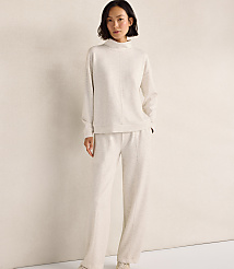 A woman wearing a cream-colored matching sweater and pants set stands against a plain, light-colored wall. carousel Product Image 1