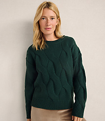 A blonde woman wearing a dark green cable-knit sweater and tan skirt stands against a plain, light-colored wall. carousel Product Image 1