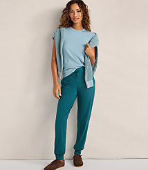 A woman wearing a light blue t-shirt, a matching cardigan, and teal joggers stands against a plain, neutral-colored wall. carousel Product Image 2