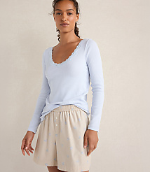 A woman wearing a light blue long-sleeved top and beige patterned shorts stands against a plain, neutral-toned wall. carousel Product Image 1