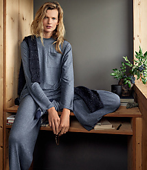 A woman in a grey lounge set and dark scarf sits on a wooden bench against a neutral grey wall with a potted plant to her side. carousel Product Image 4