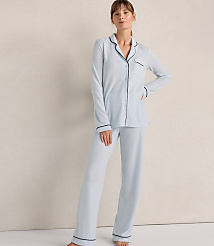 A woman wearing light blue pajamas with dark piping stands against a plain, neutral-toned wall. carousel Product Image 1