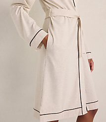 A person wears a cream-colored robe with dark piping against a plain, neutral-toned wall. carousel Product Image 2