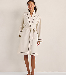 A woman wearing a cream-colored robe with dark piping stands against a plain, neutral-toned wall. carousel Product Image 1