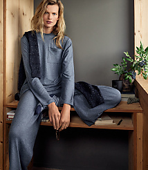 A woman in a grey lounge set sits on a wooden bench against a plain grey wall, with a small potted plant and books nearby. carousel Product Image 4