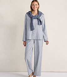 A smiling woman wearing blue and white striped pajamas with a grey sweater draped over her shoulders stands against a plain, light-colored wall. carousel Product Image 1