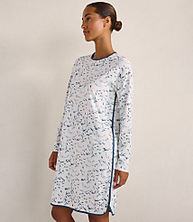 A woman wearing a white long-sleeved dress with a blue floral pattern stands against a plain, light-colored wall. carousel Product Image 2