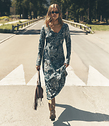 A woman wearing a long, patterned blue dress and sunglasses walks down a sunny road lined with trees and wooden railings. carousel Product Image 4