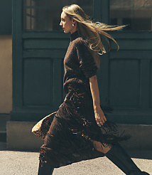 A blonde woman in a brown knit top and fringed skirt walks past a dark green paneled wall. carousel Product Image 1