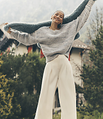 A smiling woman wearing a grey sweater and white trousers holds a blue faux-fur shawl aloft against a backdrop of a traditional house and misty mountains. carousel Product Image 4
