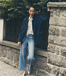 A woman wearing a blue velvet blazer and shimmering wide-leg jeans leans against a stone wall with lush green foliage in the background. carousel Product Image 1